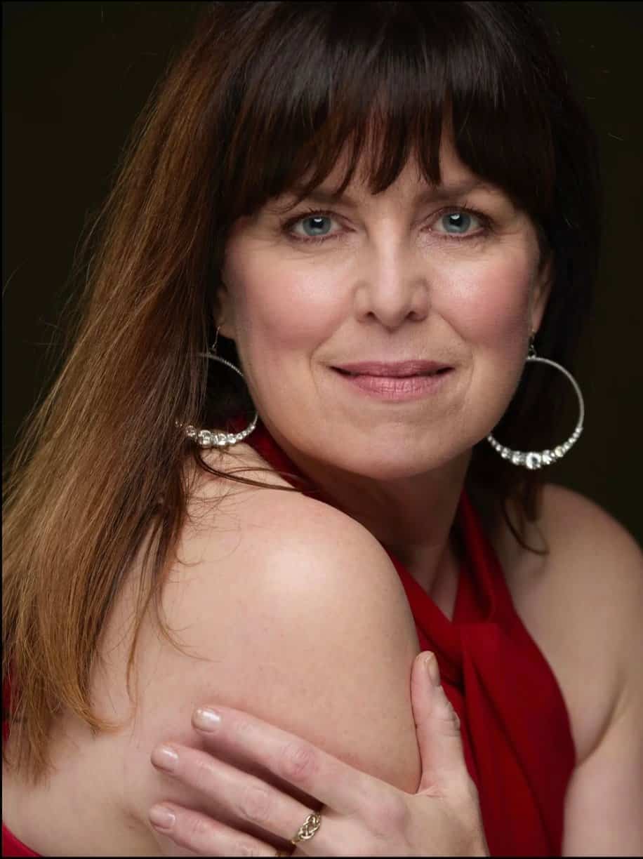 Close-up portrait of a woman with brown hair, blue eyes, wearing large hoop earrings and a red top, representing the Orpheus Pagan Chamber Choir.